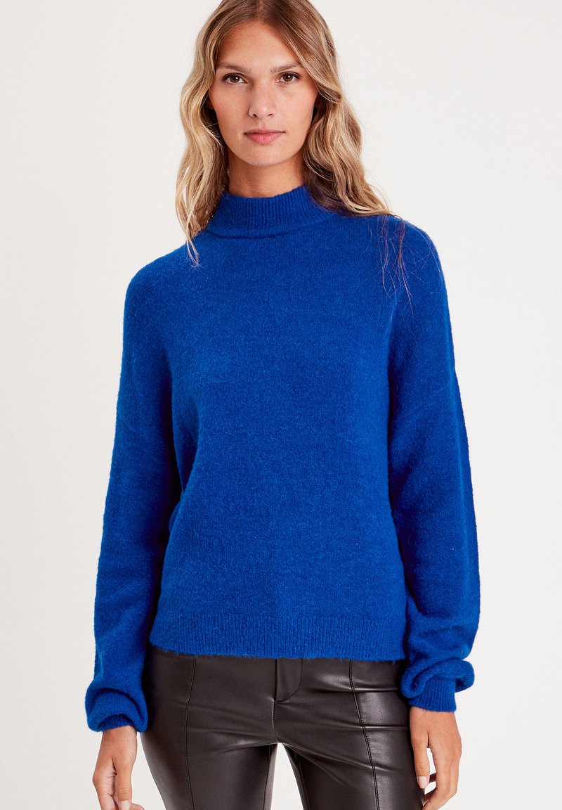 Blauer Rollkragenpullover mit einer weichen, flauschigen Textur und lässiger Passform. Hat lange Ärmel und gerippte Bündchen, kombiniert mit Lederhosen.