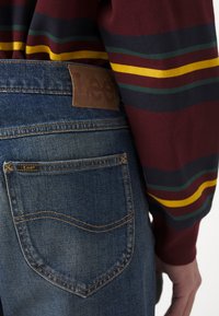 Lee džinsi ar zilo denim audumu, ar brūnu ādas etiķeti jostasvietā un aizmugurējo kabatu ar diskrētu šuvumu. Strīpota garām piedurknēm krekls.