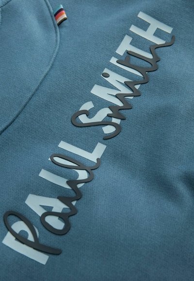 Sinine särk tekstureeritud viimistlusega, millel on silmapaistev must kiri "PAUL SMITH" korralikus stiilis ja väike mitmevärviline silt krael.