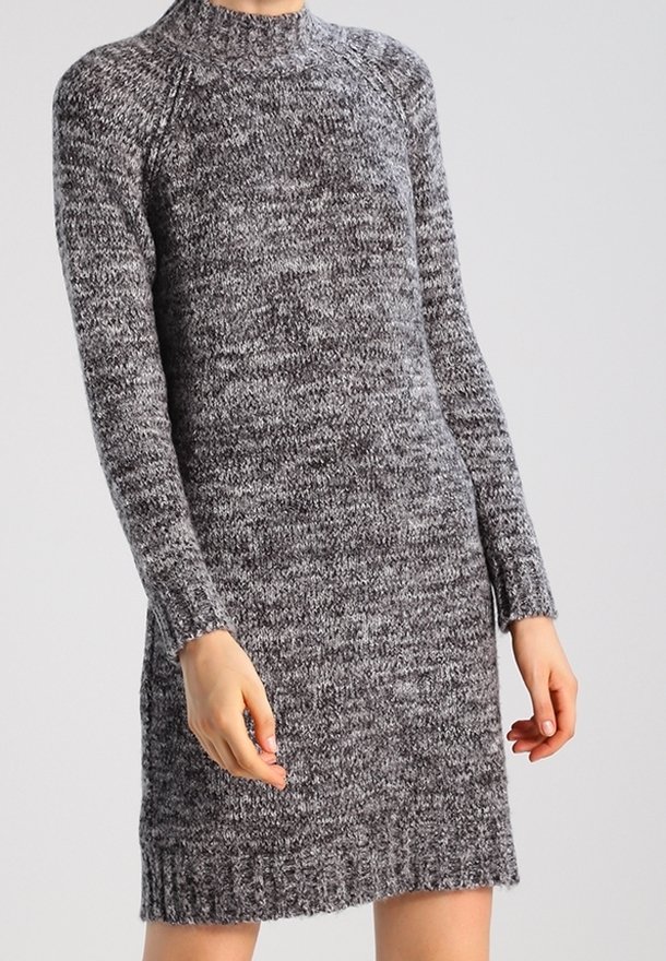 Personne portant une robe pull grise à manches longues et col montant, debout devant un fond clair uni.