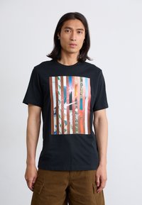 Mandlig model iført en sort t-shirt med et farverigt stribet grafisk design. Baggrunden er enkel, hvilket fokuserer på hans udtryk og tøj.