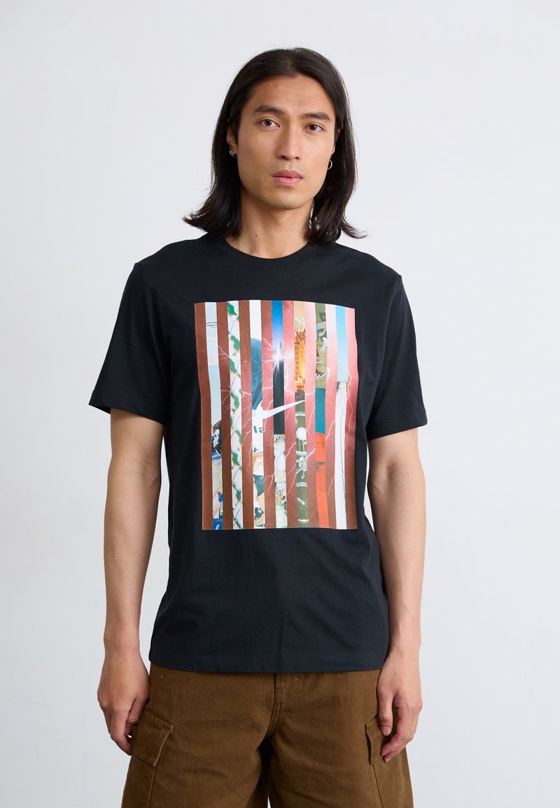 Mandlig model iført en sort t-shirt med et farverigt stribet grafisk design. Baggrunden er enkel, hvilket fokuserer på hans udtryk og tøj.