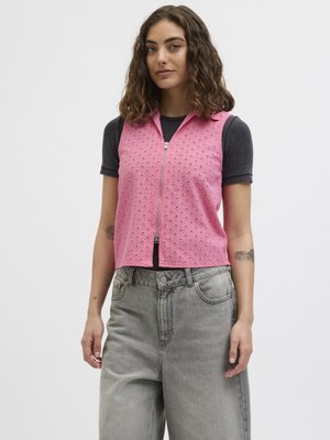 Ung kvinde med skulderlangt krøllet hår iført en pink ærmeløs vest med lynlås over en sort kortærmet skjorte og løstsiddende grå jeans.