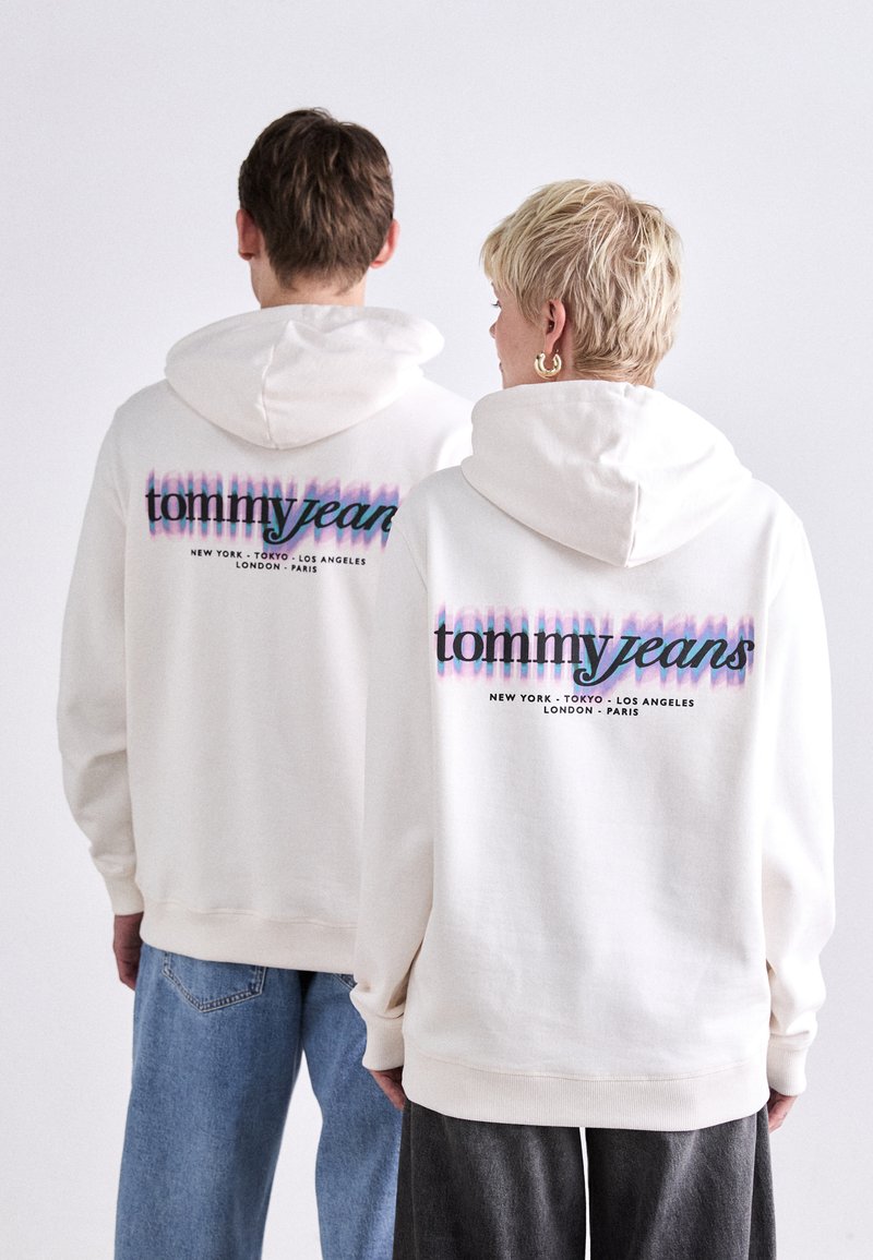 Sweatshirt à capuche blanche avec un imprimé graphique en bleu et rose portant la mention "tommyjeans" et des noms de villes. Tissu doux, coupe décontractée, poignets côtelés.