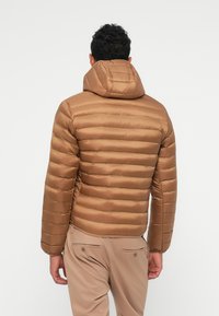 Giacca a vento marrone con design trapuntato orizzontale e cappuccio. Texture liscia e lucida; indossata sopra pantaloni beige, mostrando una silhouette elegante.