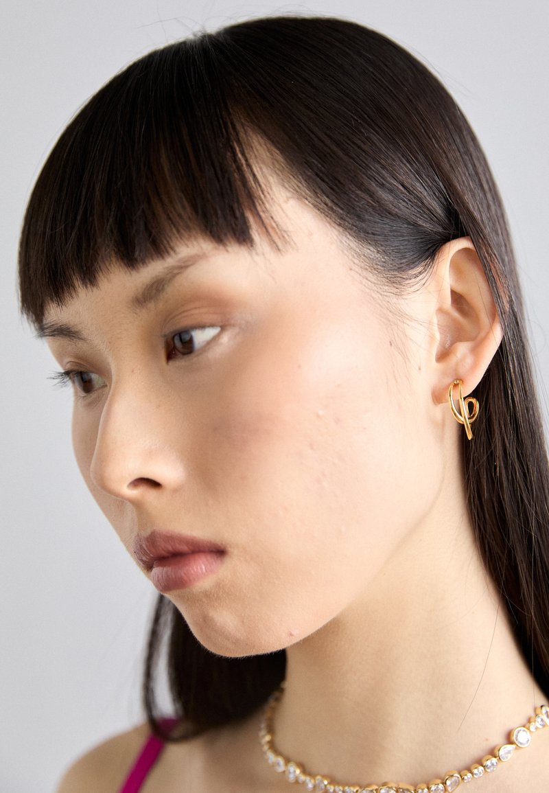 Zlatý kruhový náušník nasadený na pravom uchu, s otvoreným dizajnom. Modelka má rovné, tmavé vlasy a minimálny make-up.