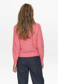 Rozā, adīta džemperis ar brīvu piegriezumu, ar garām piedurknēm un ribotām malām. Kombinēts ar tumšām denima biksēm.