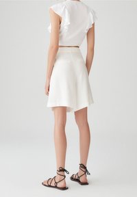 Mujer vestida con shorts blancos de cintura alta y un top corto blanco con volantes, combinado con sandalias planas negras con cordones, de pie sobre un suelo blanco.