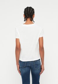 Persona con i capelli intrecciati che indossa una maglietta bianca a maniche corte e jeans di denim blu, in piedi contro uno sfondo bianco, vista posteriore.