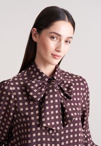 Braune Bluse mit cremefarbenem quadratischem Muster, hohem Kragen und Schleifendetail. Glattes Gewebe mit dezentem Glanz; mit Knopfverschluss und Puffärmeln.