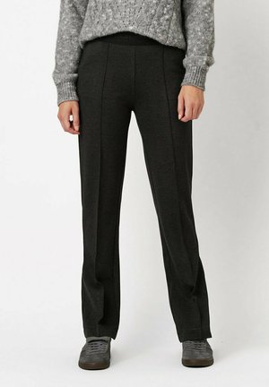 Schwarze, weite Hosen aus weichem Stoff mit einem strukturierten Bund, versehen mit Nahtdetails und einer lässigen Passform. Kombiniert mit grauem Schuhwerk.