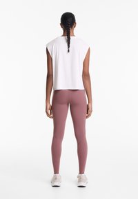 White cropped sleeveless top and mauve leggings with seam detailing; model wearing light sneakers, with hair in a braid.