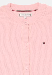 Ανοιχτό ροζ πλεκτό ζακέτα με ραβδωτή υφή, διαθέτοντας στρογγυλή λαιμόκοψη, τρία κουμπιά και μια μικρή ετικέτα λογότυπου σε κόκκινο, λευκό και μπλε χρώμα.