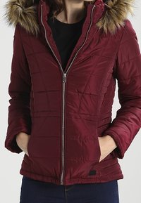 Bordeauxfarvet quiltet jakke med pelskantet hætte, lynlås foran, sidelommer og ribstrikkede manchetter, med en glat, skinnende tekstur.