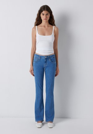 Young woman with long curly hair standing straight wearing a white tank top, blue jeans, and white shoes against a plain light background.