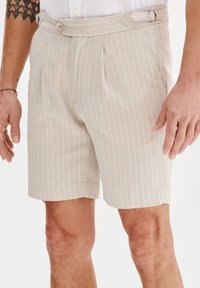 Beżowe szorty w paski wykonane z lekkiego materiału, z plisami, bocznym zapięciem na klamrę oraz klasycznym krojem dopasowanym.