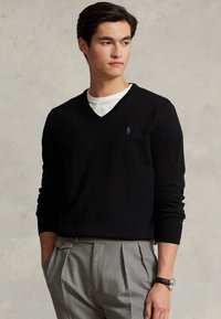 Fekete V nyakú pulóver, kötött anyagból, slim fit fazonnal, kis logóval a mellkasán. Tailored szürke nadrággal és egy órával párosítva.