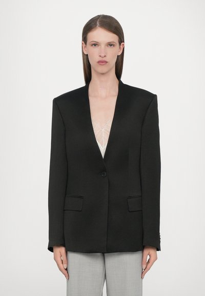 Zwarte oversized blazer met een enkele knoop, gekartelde revers, twee voorzakken en een gladde textuur, gecombineerd met een lichte onderkleding.