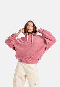 Oversized pink hoodie with a textured fabric, drawstring hood, ribbed hem, and heart logo detail. Paired with light-colored cargo pants.