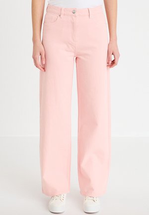 Vrouw draagt lichtroze jeans met wijde pijpen en hoge taille, met zakken aan de voorkant en witte sneakers, staand tegen een effen witte achtergrond.