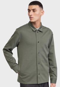 Chaqueta de color verde oliva, de manga larga y con botones, hecha de una tela suave, que cuenta con un cuello tipo solapa y botones a presión negros en la parte delantera.
