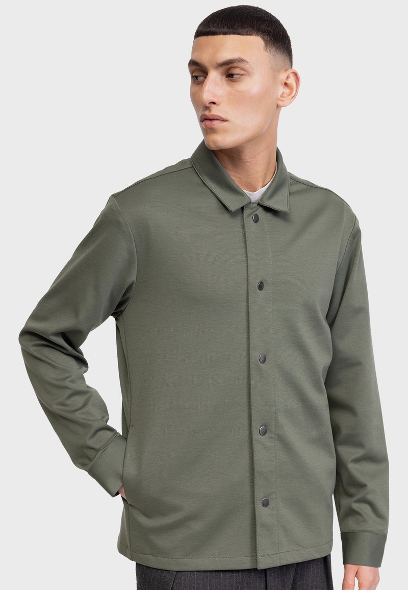 Chaqueta de color verde oliva, de manga larga y con botones, hecha de una tela suave, que cuenta con un cuello tipo solapa y botones a presión negros en la parte delantera.