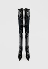 Bottes noires en cuir verni, haute cuisse, avec un bout pointu, des détails de fermeture éclair verticaux, une texture lisse et brillante, et un design structuré.