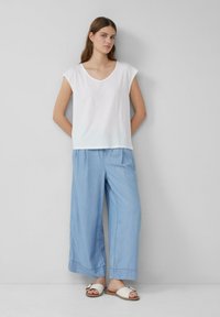 Weißes V-Ausschnitt-T-Shirt mit kurzen Ärmeln, kombiniert mit hellblauen, weit geschnittenen Hosen und weißen Sandalen mit goldenen Akzenten. Glatte Stoffstruktur.