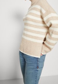 Osoba w beżowym i białym swetrze w paski z bocznymi rozporkami oraz niebieskich dżinsach, z jedną ręką w kieszeni, na jednolitym tle.