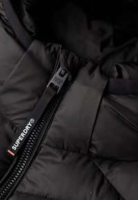 Manteau rembourré noir avec un extérieur lisse et brillant, doté d'un matelassage et d'un tirette de fermeture éclair à la marque Superdry.