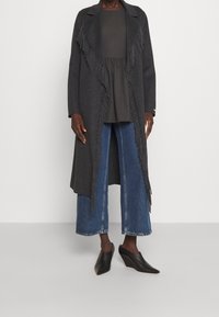 Grå franskantskåret frakke over en mørk top, parret med brede blå jeans og sorte kilehæle. Glat tekstur, lang silhuet.