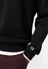 Felpa nera con orlo e polsini a coste, caratterizzata da un piccolo logo ricamato sulla manica. Tessuto morbido, indossata sopra una camicia bianca.