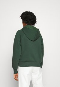 Felpa verde scuro con tasca frontale, polsini e orlo a coste. Presenta un cappuccio e un tessuto morbido e strutturato. Indossata con pantaloni di colore chiaro.