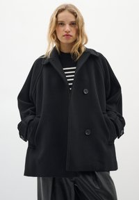 Black oversized wool coat with a notched collar, double buttons, and patch pockets. Worn over a striped black and white top. Smooth texture.