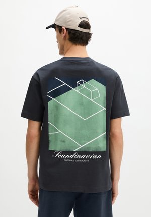 Mann trägt ein schwarzes T-Shirt mit grüner und marineblauer Fußballfeld-Grafik und dem Schriftzug "Scandinavian Football Community" auf dem Rücken.