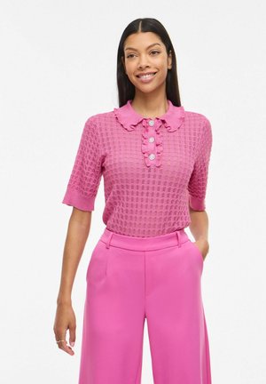 Femme souriante, portant un haut texturé rose à manches courtes avec un col à volant et des boutons, associé à un pantalon rose taille haute.