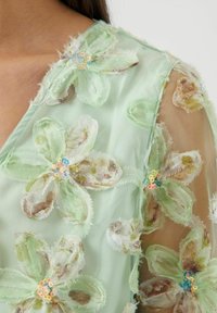 Lys grønn mesh-stoff dekorert med lagdelte blomsterapplikasjoner i varierende nyanser. Hver blomst har paljetter i midten for ekstra detaljer.