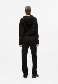 Persona con capelli intrecciati che indossa un hoodie nero, pantaloni neri e scarpe nere, in piedi con le spalle rivolte verso l'osservatore su uno sfondo bianco.