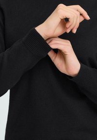 Schwarzer Strickpullover mit gerippten Bündchen, glatter Textur, tailliertem Design und Nahaufnahme von Händen, die den Ärmel anpassen.