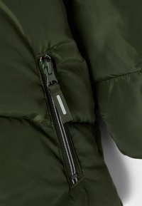 Sezione di giacca imbottita verde oliva con zip verticale e linguetta in tessuto, texture trapuntata e superficie liscia e lucida.