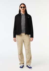 Chaqueta negra con cuello, camiseta polo gris, pantalones cargo beige y zapatillas deportivas azules, con tejido texturizado y bolsillos en los pantalones.