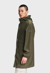 Homme portant un long imperméable vert olive avec capuche, debout de côté devant un fond clair uni.