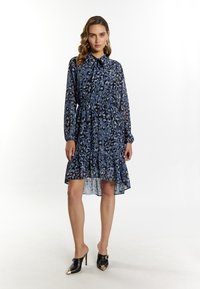 Blauwe luipaardprint jurk met lange mouwen, knoopsluiting, elastische taille en ruches aan de onderkant. Gedragen met zwarte open teen hakken.