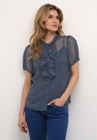 Blusa blu trasparente con dettagli arricciati sul davanti, maniche a sbuffo corte e chiusura a bottoni, indossata con jeans blu su uno sfondo neutro.