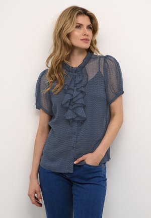 Blå gennemsigtig bluse med volangdetalje foran, korte pufærmer og knaplukning, båret med blå jeans mod en neutral baggrund.