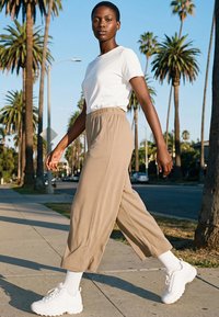 Nő sétál a pálmafákkal szegélyezett járdán, fehér pólót, bézs színű bő szárú nadrágot, fehér zoknit és masszív fehér sportcipőt visel.