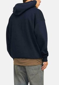 Sudadera azul marino con un corte relajado, tela texturizada y una gran capucha. El modelo la combina con una camisa beige y unos vaqueros gris claro.