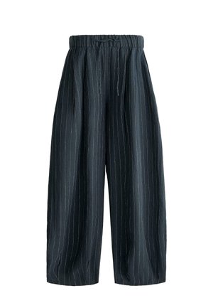 Vrouw staat met handen in de zakken, draagt een witte mouwloze cropped top, losse donkere broek met fijne strepen en zwarte sandalen op een betegelde vloer.