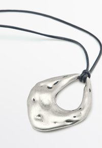 Pendente in metallo argentato con una forma organica e astratta e un taglio centrale, appeso a un cordino nero. Presenta una texture irregolare e dettagli decorativi.