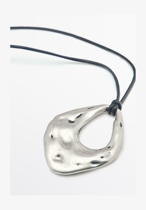 Srebrny medalion o metalicznym wykończeniu, w organicznym, abstrakcyjnym kształcie z centralnym wycięciem, zawieszony na czarnym sznurku. Posiada nierówną fakturę i akcenty.